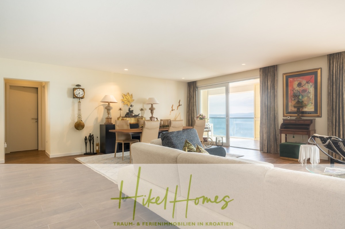 Helles, modernes Wohnzimmer mit elegantem Dekor, großen Fenstern mit Blick auf das Meer, hellen Holzböden, stilvollen Möbeln, Wandkunst, einem Klavier und einer Schiebetür, die auf einen Balkon führt. Der Text "Hikel Homes" ist am unteren Rand eingeblendet. - MEERBLICK & KUNST Designer Luxus Appartement mit 3 Schlafzimmer – großer Loggia