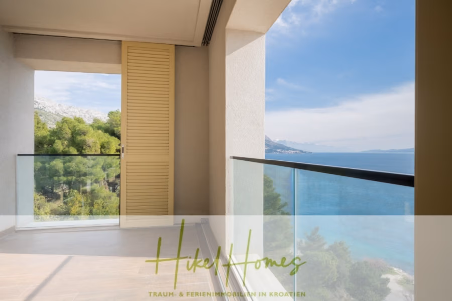 MEERBLICK & KUNST Designer Luxus Appartement mit 3 Schlafzimmer – großer Loggia - Ein geräumiger Balkon mit Glasgeländer bietet einen malerischen Blick auf das Meer und die üppig grünen Hügel. Der Balkon hat helle Wände und eine offene Tür mit Fensterladen. Der Boden ist gefliest und schafft eine ruhige Atmosphäre. (41081)