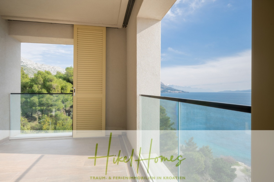 MEERBLICK & KUNST Designer Luxus Appartement mit 3 Schlafzimmer – großer Loggia - Ein Balkon mit Glasgeländer bietet einen Blick auf das blaue Meer und die grünen Bäume, im Hintergrund sind die Berge zu sehen. Eine teilweise geöffnete gelbe Rollladentür führt ins Innere. Der Schriftzug "Hikel Homes" erscheint am unteren Rand. (45385)