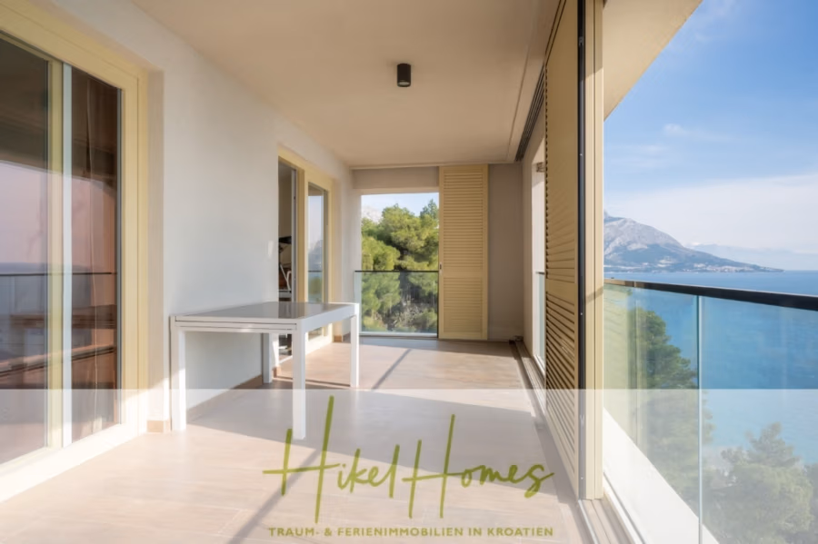 MEERBLICK & KUNST Designer Luxus Appartement mit 3 Schlafzimmer – großer Loggia - Ein geräumiger Balkon mit Glasgeländer bietet einen malerischen Blick auf das Meer und die Berge in der Ferne. Ein weißer Tisch steht an der Wand. Der Bereich ist schattig, mit beigefarbenen Fliesen und cremefarbenen Wänden, was eine ruhige Atmosphäre schafft. (41082)