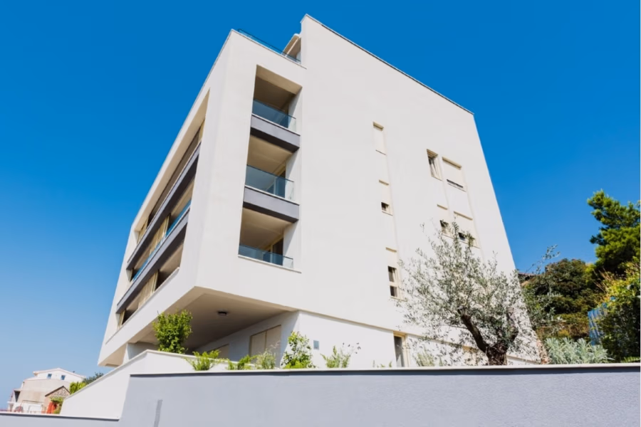 MEERBLICK & KUNST Designer Luxus Appartement mit 3 Schlafzimmer – großer Loggia - Ein modernes, beigefarbenes, mehrstöckiges Wohnhaus mit Balkonen und großen Fenstern vor einem strahlend blauen Himmel. Im Vordergrund zieren grüne Sträucher und Bäume die Stadtlandschaft und verleihen ihr einen Hauch von Natur. (41093)