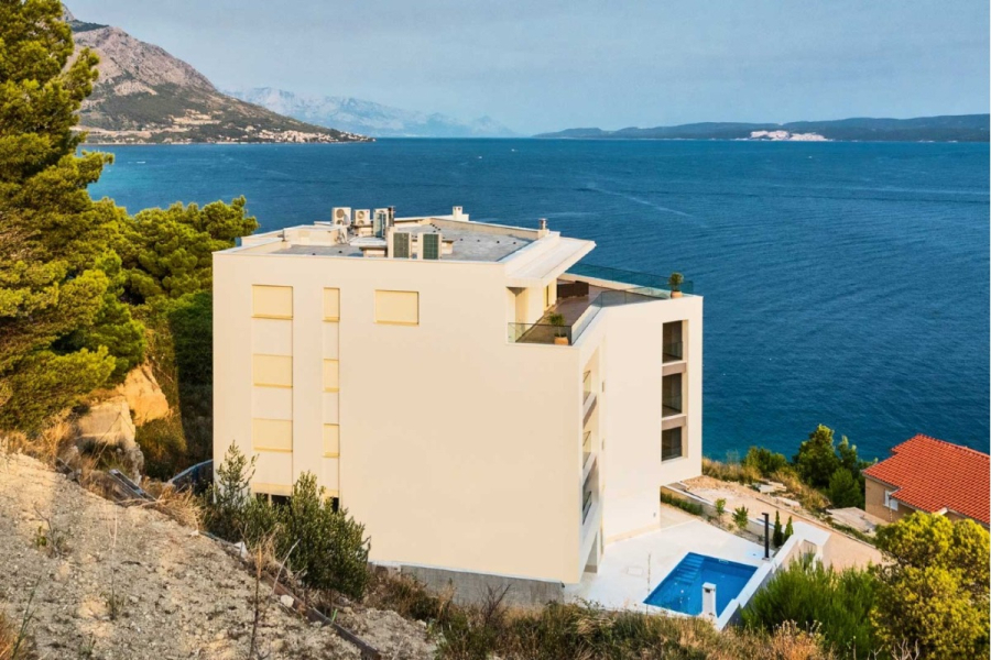 MEERBLICK & KUNST Designer Luxus Appartement mit 3 Schlafzimmer – großer Loggia - Ein modernes weißes Gebäude mit flachem Dach steht auf einem Hügel mit Blick auf ein tiefblaues Meer, mit Bergen und mehr Wasser in der Ferne. Hohe Kiefern säumen die Szene, und unten ist ein Pool zu sehen. (45399)