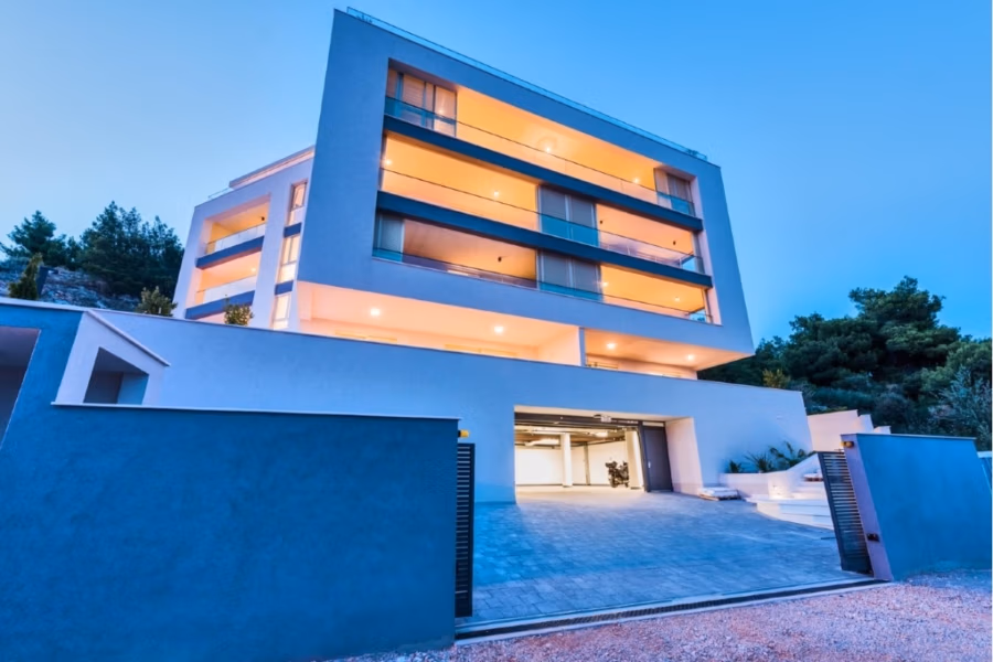 MEERBLICK & KUNST Designer Luxus Appartement mit 3 Schlafzimmer – großer Loggia - Ein modernes, mehrstöckiges Gebäude mit minimalistischem Design verfügt über große Balkone und eine warme Innenbeleuchtung. Es steht vor einem Abendhimmel, umgeben von Bäumen, mit einer offenen Garage und einer gepflasterten Auffahrt im Vordergrund. (41094)