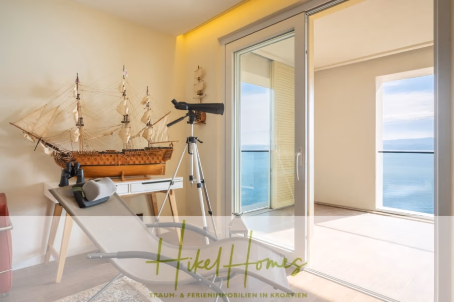 MEERBLICK & KUNST Designer Luxus Appartement mit 3 Schlafzimmer – großer Loggia - Ein Zimmer mit Meerblick, einem großen Schiffsmodell auf einem Tisch, einem Teleskop und einem Liegestuhl gegenüber einer Glasschiebetür, die auf einen Balkon führt. Unten ist der Schriftzug „Hikel Homes“ eingeblendet. (41080)