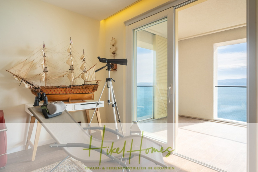 MEERBLICK & KUNST Designer Luxus Appartement mit 3 Schlafzimmer – großer Loggia - Ein helles Küstenzimmer mit einem Loungesessel, einem Teleskop und einem großen Schiffsmodell neben Glastüren, die zu einem Balkon mit Blick auf das Meer führen. Das "Hikel Homes"-Logo und die Textüberlagerung befinden sich am unteren Rand. (45383)