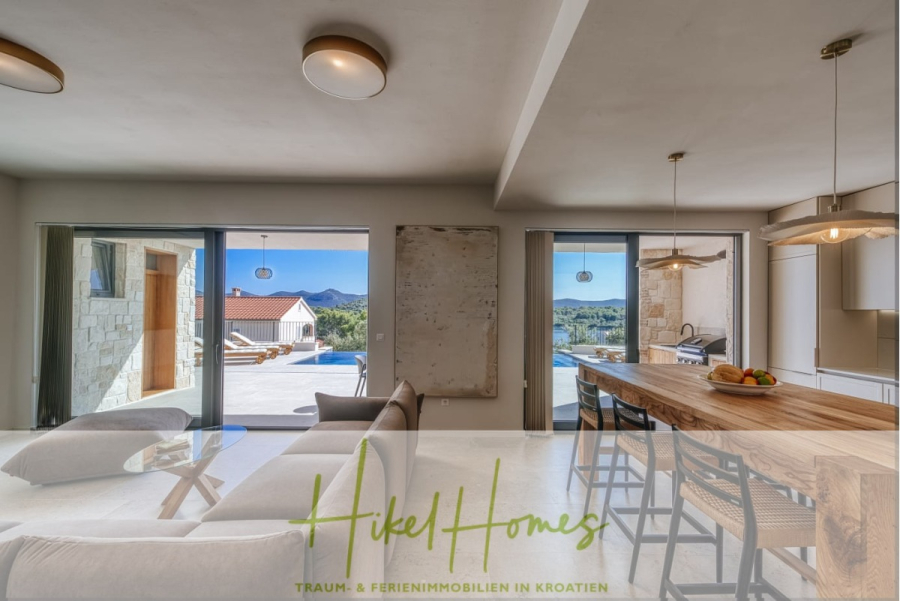 Moderne Meerblick Villa mit Infinity-Pool, Bootsanleger, 100m zum Meer - Moderner, heller Wohn- und Essbereich mit bodentiefen Fenstern und Blick auf einen Pool, die Berge und ein Haus. Es gibt eine Kücheninsel mit Hockern, ein Sofa und natürliches Licht, das den stilvollen Raum erfüllt. (44433)