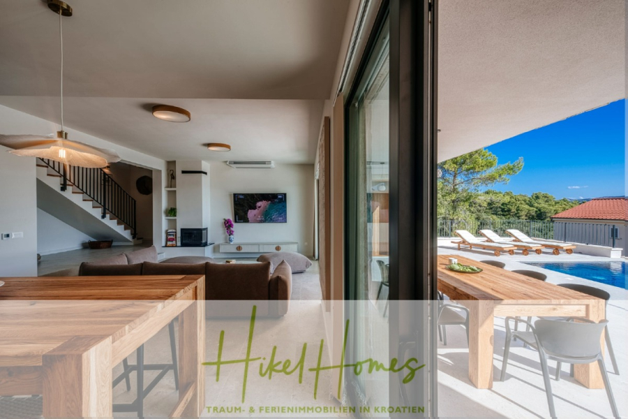 Moderne Meerblick Villa mit Infinity-Pool, Bootsanleger, 100m zum Meer - Moderner, offener Wohn- und Essbereich mit Holzmöbeln, einem Fernseher und großen Fenstern mit Blick auf eine Terrasse mit einem Pool, Sonnenliegen und einer malerischen Aussicht. Das Logo und der Slogan von "Hikel Homes" erscheinen am unteren Rand des Bildes. (44431)