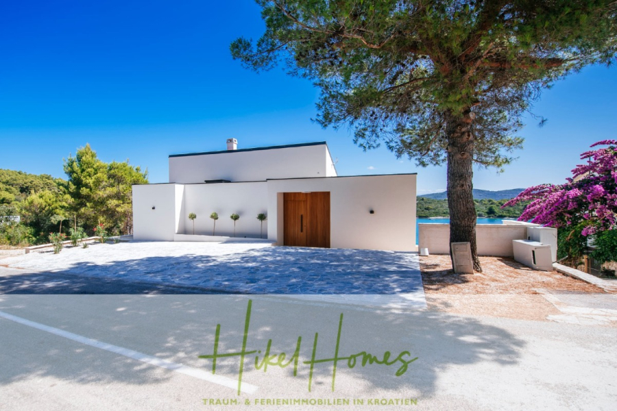 Moderne Meerblick Villa mit Infinity-Pool, Bootsanleger, 100m zum Meer - Moderne weiße Villa mit Holztür, Flachdach und minimalistischer Landschaftsgestaltung, vor blauem Himmel und umgeben von Bäumen und Grünflächen. Im Hintergrund ist das Meer zu sehen. Der Text lautet "Hikel Homes". (44455)