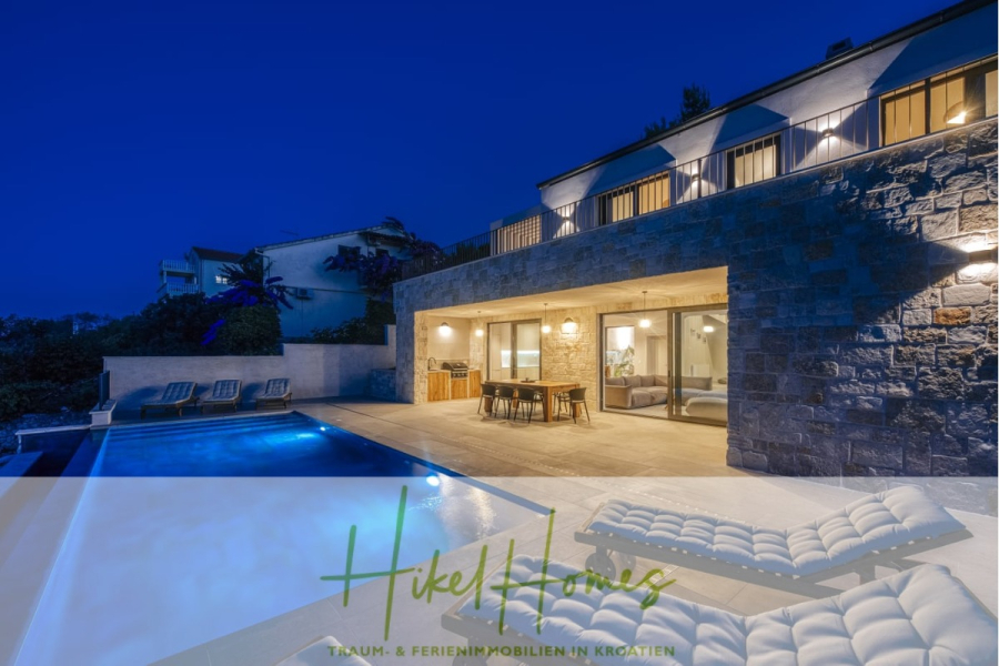 Moderne Meerblick Villa mit Infinity-Pool, Bootsanleger, 100m zum Meer - Moderne Steinvilla bei Nacht mit beleuchtetem Pool, Liegestühlen und Essbereich im Freien. Große Glastüren geben den Blick auf einen warm beleuchteten Innenraum frei. Das Logo und der Slogan von "Hikel Homes" erscheinen am unteren Rand des Bildes. (44453)