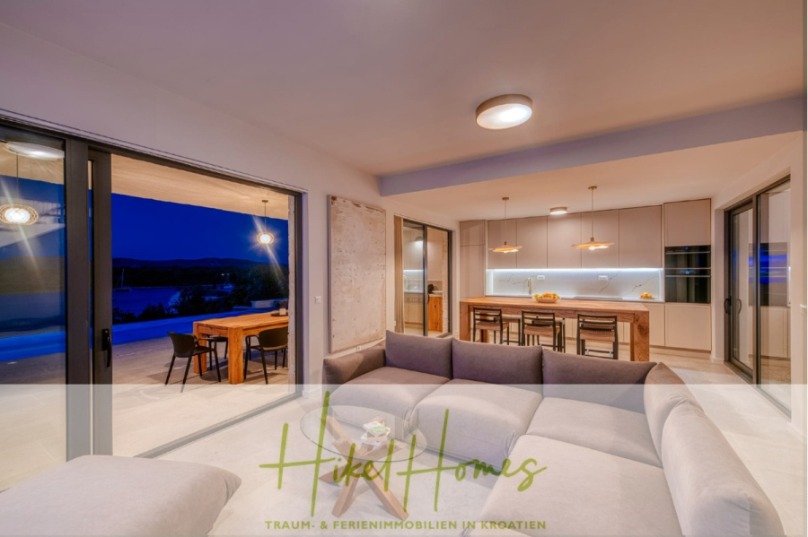 Moderne Meerblick Villa mit Infinity-Pool, Bootsanleger, 100m zum Meer - Modernes offenes Wohnzimmer und Küche mit neutralen Farbtönen, einem großen Sektionssofa und Glasschiebetüren, die in der Abenddämmerung zu einem Essbereich im Freien führen. Der Schriftzug "Hikel Homes" erscheint am unteren Rand. (44454)