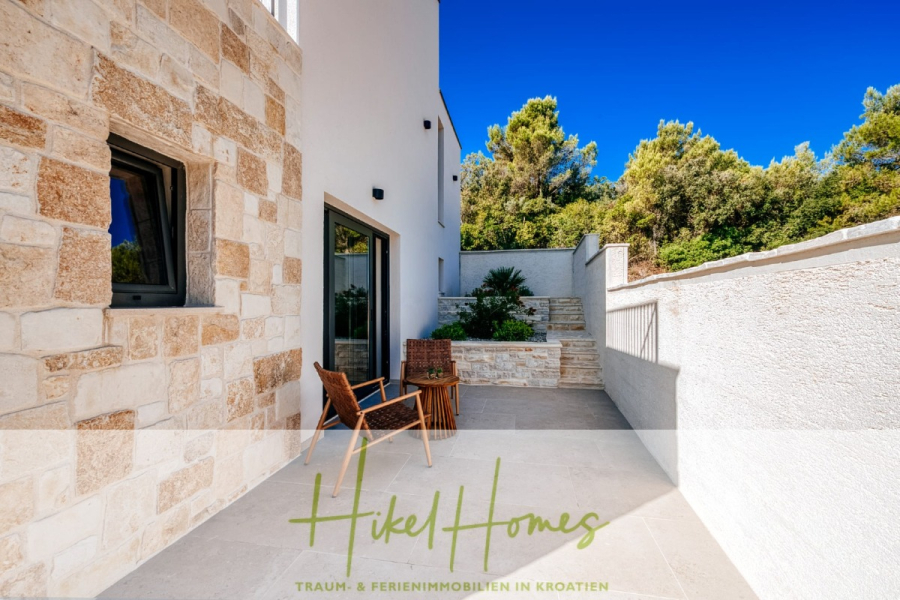 Moderne Meerblick Villa mit Infinity-Pool, Bootsanleger, 100m zum Meer - Moderne Steinterrasse mit einem Korbstuhl, einem kleinen Tisch und üppigem Grün vor einem weißen Steinhaus unter einem klaren blauen Himmel. Der Text lautet: "Hikel Homes. Traum- & Ferienimmobilien in Kroatien.". (44438)
