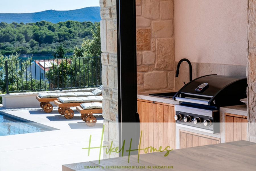 Moderne Meerblick Villa mit Infinity-Pool, Bootsanleger, 100m zum Meer - Außenküche mit eingebautem Grill und Spüle, mit Blick auf den Pool und die Liegestühle. Im Hintergrund sind grüne Bäume und Hügel unter einem klaren Himmel zu sehen. Die Szene suggeriert eine entspannende Urlaubsumgebung. (44437)
