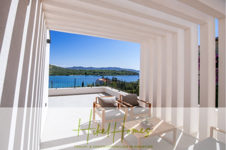 Moderne Meerblick Villa mit Infinity-Pool, Bootsanleger, 100m zum Meer - Eine moderne Terrasse mit zwei Holzstühlen und einem kleinen Tisch bietet einen Blick auf einen blauen See und grüne Hügel unter einem klaren Himmel. Weiße Pergola-Balken werfen Schatten auf den hellen Terrassenboden. (44443)
