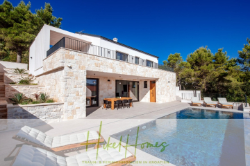 Moderne Steinvilla mit großen Fenstern, Essbereich im Freien, Pool und Sonnenliegen, umgeben von Pinienbäumen unter einem klaren blauen Himmel. Logo und Text von "Hikel Homes" erscheinen am unteren Rand des Bildes. - Moderne Meerblick Villa mit Infinity-Pool, Bootsanleger, 100m zum Meer