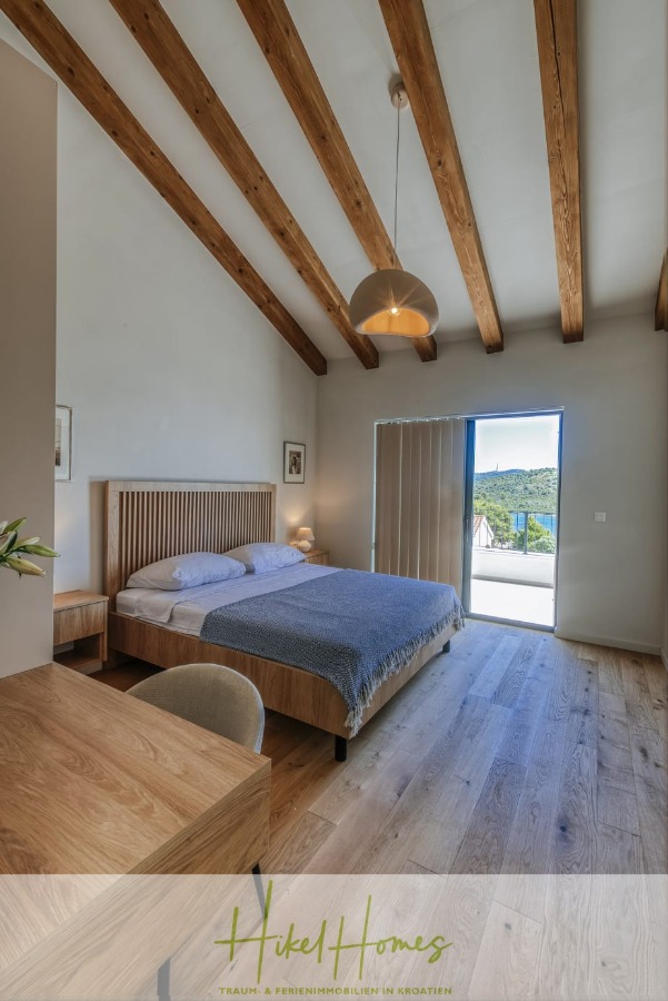 Moderne Meerblick Villa mit Infinity-Pool, Bootsanleger, 100m zum Meer - Ein modernes Schlafzimmer mit Holzbalken, einem Doppelbett mit blauer Bettwäsche, einer Hängelampe, einem Holztisch mit Stuhl und einer Glastür, die auf einen Balkon mit Panoramablick führt. (44452)