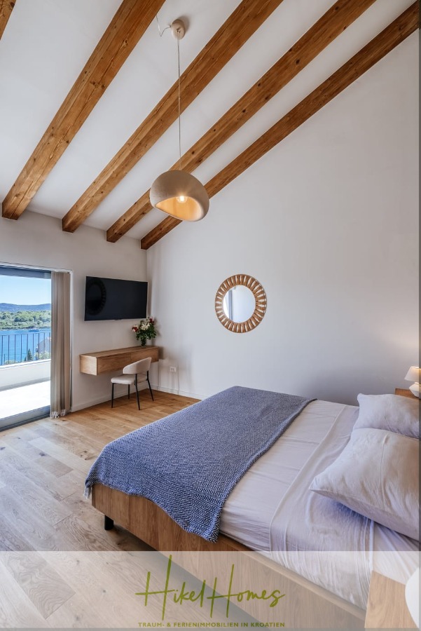 Moderne Meerblick Villa mit Infinity-Pool, Bootsanleger, 100m zum Meer - Ein modernes Schlafzimmer mit Holzbalken, einem Doppelbett mit grauer Decke, einem an der Wand montierten Fernseher, einem Schreibtisch mit Stuhl, einem runden Spiegel und Schiebetüren, die zu einem Balkon mit Meerblick führen. (44448)