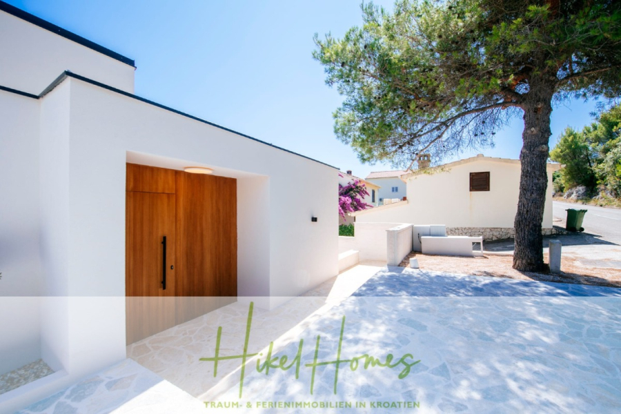 Moderne Meerblick Villa mit Infinity-Pool, Bootsanleger, 100m zum Meer - Modernes weißes Haus mit Holztür, Steinauffahrt und einem großen Baum im Vorgarten. Die Szene ist sonnig mit blauem Himmel. Der Text am unteren Rand lautet: "Hikel Homes - Traum- & Ferienimmobilien in Kroatien. (44457)