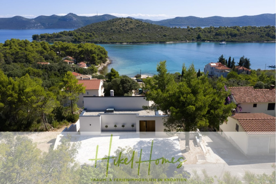 Moderne Meerblick Villa mit Infinity-Pool, Bootsanleger, 100m zum Meer - Modernes weißes Haus, umgeben von Bäumen, mit Blick auf eine ruhige blaue Bucht mit grünen Hügeln im Hintergrund. In der Nähe befinden sich Häuser mit roten Dächern. Das Bild enthält das Logo "Hikel Homes" am unteren Rand. (44430)