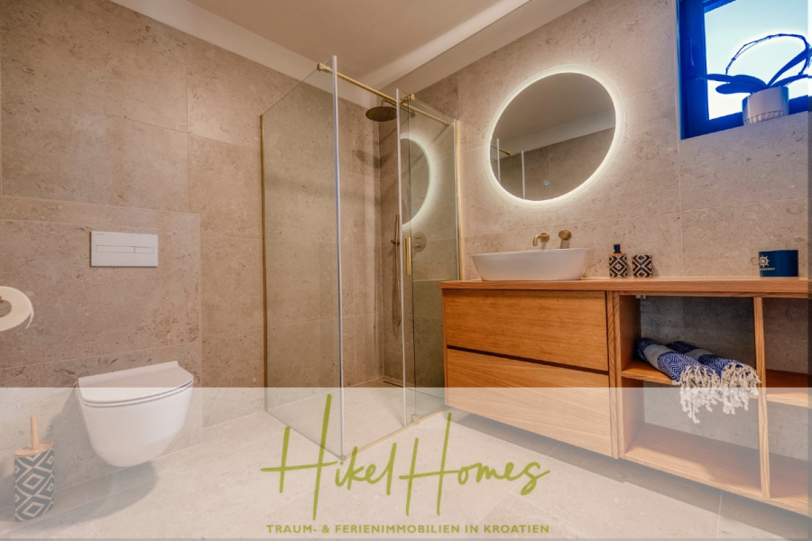 Moderne Meerblick Villa mit Infinity-Pool, Bootsanleger, 100m zum Meer - Modernes Badezimmer mit beigen Fliesen, einer gläsernen Dusche, einem runden, hinterleuchteten Spiegel, einem hölzernen Waschtisch mit Gefäßwaschbecken, offenen Regalen mit Handtüchern, einem kleinen Fenster und einer getopften Orchidee auf dem Tresen. (44444)