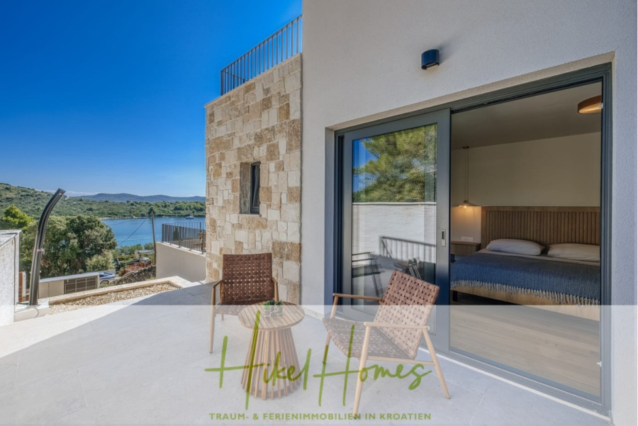Moderne Meerblick Villa mit Infinity-Pool, Bootsanleger, 100m zum Meer - Moderne Terrasse mit zwei Stühlen und einem Tisch vor einem weißen Steinhaus, mit Blick auf einen malerischen See und grüne Hügel; Schlafzimmer mit Doppelbett durch große Glasschiebetür sichtbar. (44442)