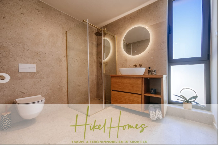 Moderne Meerblick Villa mit Infinity-Pool, Bootsanleger, 100m zum Meer - Modernes Badezimmer mit beigen Steinwänden, einer gläsernen Dusche, einem runden beleuchteten Spiegel über einem hölzernen Waschbecken mit Gefäß, einer Wandtoilette und einem Fenster mit einer Topfpflanze daneben. (44449)