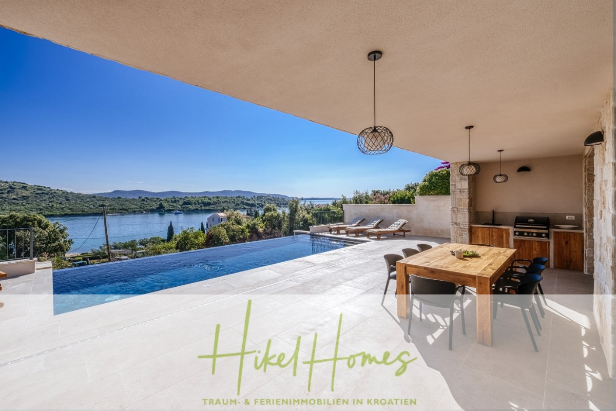 Eine moderne überdachte Terrasse mit einem Esstisch, Liegestühlen und einem Grill bietet einen Blick auf einen Infinity-Pool und einen malerischen Fluss, der von grünen Hügeln unter einem klaren blauen Himmel umgeben ist. Der Text lautet "Hikel Homes" und "Traum- & Ferienimmobilien in Kroatien". - Moderne Meerblick Villa mit Infinity-Pool, Bootsanleger, 100m zum Meer