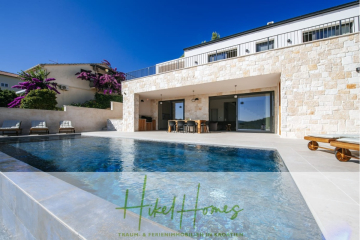 Moderne Villa mit einer Steinfassade, großen Fenstern und einem Sitzbereich im Freien neben einem klaren Swimmingpool, umgeben von Grün und lila Blumen unter einem klaren blauen Himmel. Das Logo "Hikel Homes" erscheint unten. - Moderne Meerblick Villa mit Infinity-Pool, Bootsanleger, 100m zum Meer
