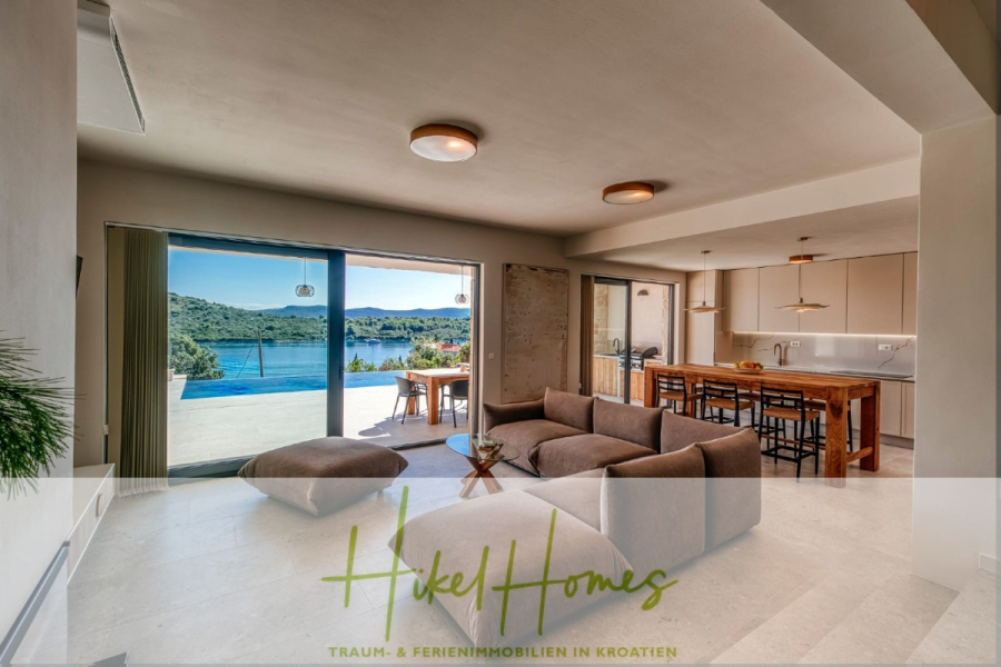 Moderne Meerblick Villa mit Infinity-Pool, Bootsanleger, 100m zum Meer - Offenes Wohnzimmer mit einem braunen Sektionssofa, moderner Küche und großen Schiebetüren, die auf eine Terrasse mit Pool und Seeblick führen. Der Schriftzug "Hikel Homes" ist am unteren Rand des Bildes zu sehen. (44434)