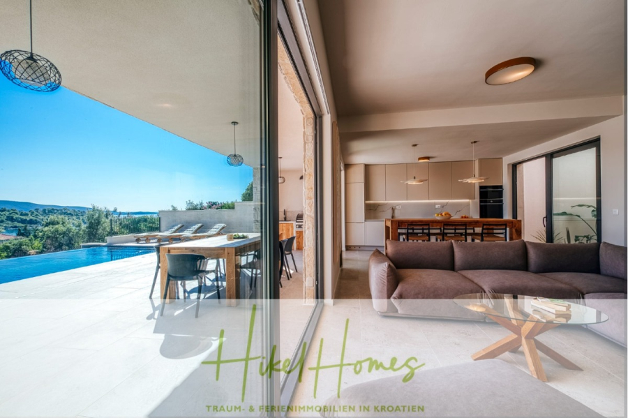 Moderne Meerblick Villa mit Infinity-Pool, Bootsanleger, 100m zum Meer - Modernes, offenes Wohnzimmer und Küche mit Glasschiebetüren, die zu einer geräumigen Terrasse, einem Essbereich im Freien und einem Infinity-Pool mit Blick auf sanfte Hügel und den blauen Himmel führen. Text überlagert: "Heikel Homes. (44436)