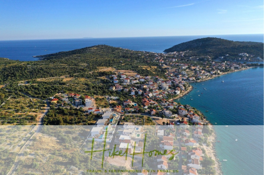Seeview 2 von 3 | Möbilierte Luxus-Meerblick Villa mit 4 SZ, Pool, Sauna, Fitness, Jacuzzi, - Luftaufnahme einer Küstenstadt in Kroatien mit Häusern entlang der Küste und grünen Hügeln mit Blick auf das blaue Meer und eine nahe gelegene Insel. Am unteren Rand des Bildes sind das HikeHomes-Logo und ein deutscher Text zu sehen. (45601)
