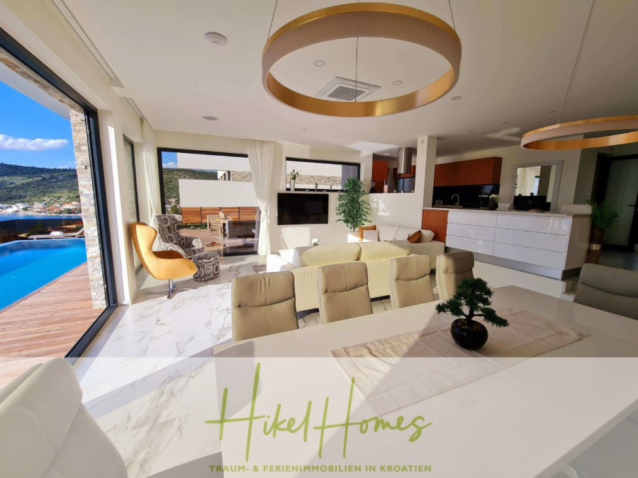Seeview 2 von 3 | Möbilierte Luxus-Meerblick Villa mit 4 SZ, Pool, Sauna, Fitness, Jacuzzi, - Moderner, offener Wohnbereich mit bodentiefen Fenstern, weißem und hölzernem Dekor, Ess- und Loungebereich, Außenpool und Blick auf die Hügel. Das Logo am Boden lautet "Hikel Homes - Traum- & Ferienimmobilien in Kroatien". (45607)
