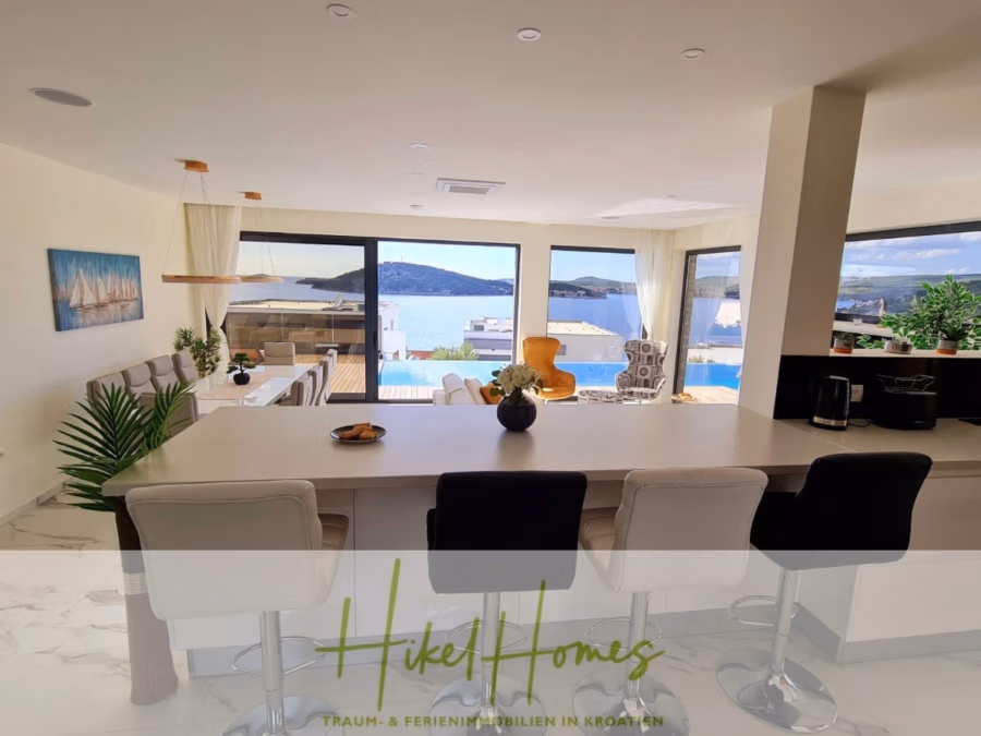 Seeview 2 von 3 | Möbilierte Luxus-Meerblick Villa mit 4 SZ, Pool, Sauna, Fitness, Jacuzzi, - Moderner, offener Wohnbereich mit Küche und Essbereich. Große Fenster bieten Blick auf Meer und Hügel. Eine Kücheninsel mit Barhockern, Pflanzen und einem Wandbehang steht auf dem Tisch. Der Text unten lautet: „Hike! Homes – Traum- & Ferienimmobilien in Kroatien.“. (41760)