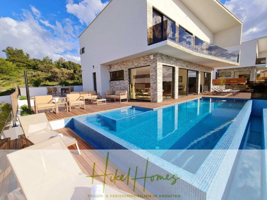 Seeview 2 von 3 | Möbilierte Luxus-Meerblick Villa mit 4 SZ, Pool, Sauna, Fitness, Jacuzzi, - Moderne weiße Villa mit großen Fenstern, einem Holzdeck, Sitzgelegenheiten im Freien und einem von Liegestühlen umgebenen Infinity-Pool. Üppiges Grün und blauer Himmel im Hintergrund. Logo und Text-Overlay am unteren Rand des Bildes. (45614)