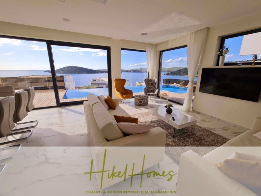 Seeview 2 von 3 | Möbilierte Luxus-Meerblick Villa mit 4 SZ, Pool, Sauna, Fitness, Jacuzzi, - Helles Wohnzimmer mit großen Fenstern und Blick auf das Meer und die Hügel. Zur Ausstattung gehören ein beiges Sofa, Sessel, ein Wandfernseher und ein Kamin. Draußen gibt es einen Pool auf der Holzterrasse. Ein Schild mit der Aufschrift „Hikel Homes“. (41757)