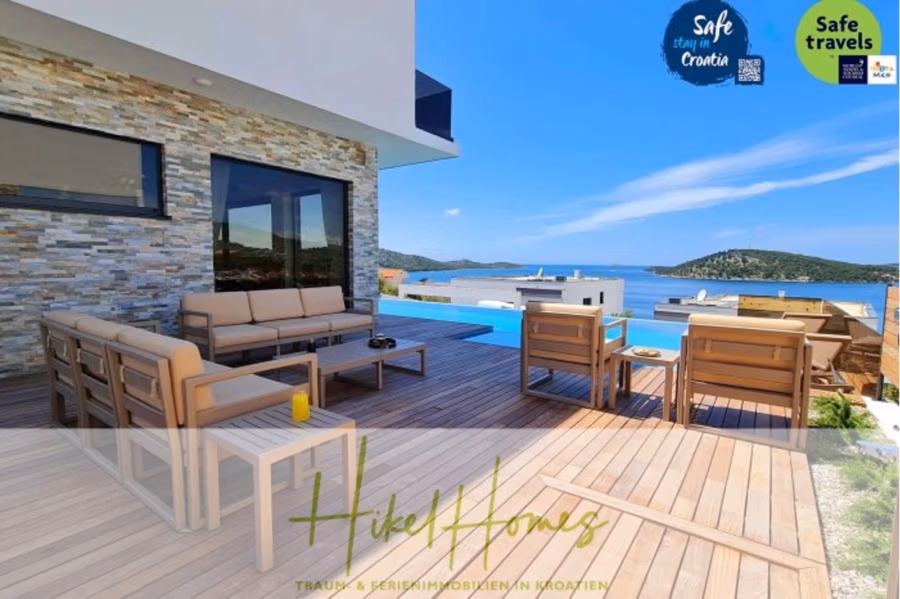 Seeview 2 von 3 | Möbilierte Luxus-Meerblick Villa mit 4 SZ, Pool, Sauna, Fitness, Jacuzzi, - Außenterrasse mit Meerblick, Holzbelag, beigefarbenen Sitzkissen, einem Glastisch und einem Pool. Steinmauern und Blick auf Hügel und Meer unter strahlend blauem Himmel. Schilder weisen auf „Sicherer Aufenthalt in Kroatien“ und „Gute Reise“ hin. (41743)