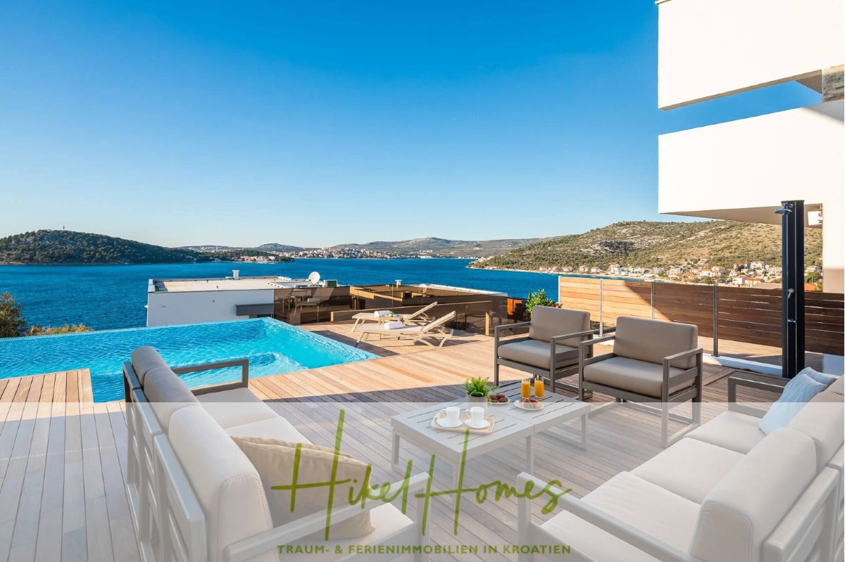 Moderne Außenterrasse mit weißen Sofas und Stühlen, einem Holzdeck, Infinity-Pool und atemberaubendem Meerblick. Im Hintergrund sind unter einem klaren blauen Himmel Hügel und Häuser an der Küste zu sehen. Text "Hikel Homes" am unteren Rand. - Seeview 2 von 3 | Möbilierte Luxus-Meerblick Villa mit 4 SZ, Pool, Sauna, Fitness, Jacuzzi,