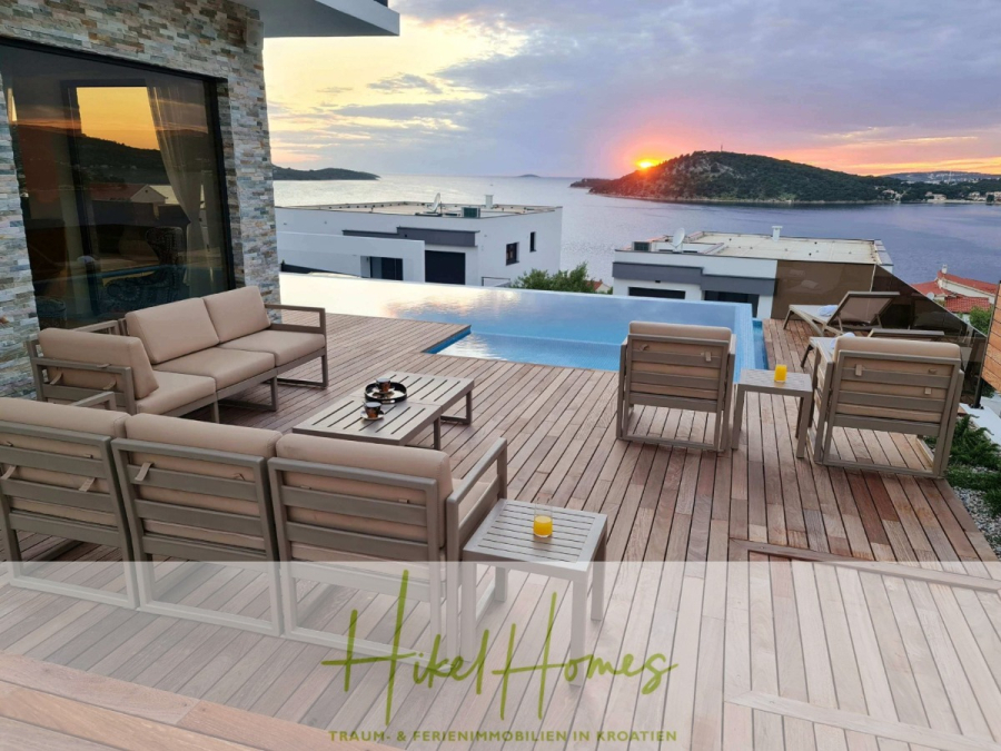 Seeview 2 von 3 | Möbilierte Luxus-Meerblick Villa mit 4 SZ, Pool, Sauna, Fitness, Jacuzzi, - Moderne Terrasse mit beigen Gartenmöbeln und kleinem Pool mit Blick auf das Meer und die Hügel bei Sonnenuntergang. Am Horizont sind nahe gelegene Häuser und eine leuchtend orangefarbene Sonne zu sehen. Das Logo "Hikel Homes" erscheint am unteren Rand des Bildes. (45605)