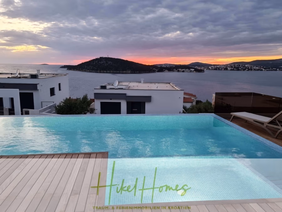 Seeview 2 von 3 | Möbilierte Luxus-Meerblick Villa mit 4 SZ, Pool, Sauna, Fitness, Jacuzzi, - Infinity-Pool mit Blick auf die Küste bei Sonnenuntergang, mit modernen weißen Gebäuden und Inseln in der Ferne. Der Himmel ist bewölkt und unten erscheint der Text „Hikel Homes“. (41748)