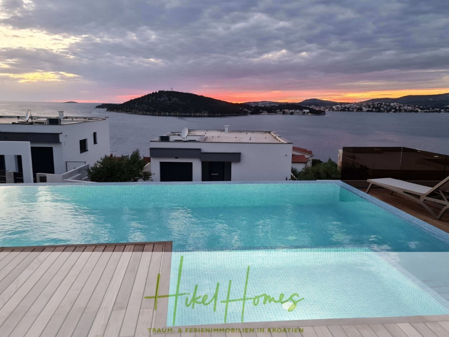 Seeview 2 von 3 | Möbilierte Luxus-Meerblick Villa mit 4 SZ, Pool, Sauna, Fitness, Jacuzzi, - Infinity-Pool mit Blick auf das Meer bei Sonnenuntergang, mit modernen weißen Gebäuden, fernen Hügeln und dramatischen Wolken. Das Logo "Hikel Homes" ist auf dem Holzdeck im Vordergrund zu sehen. (45597)