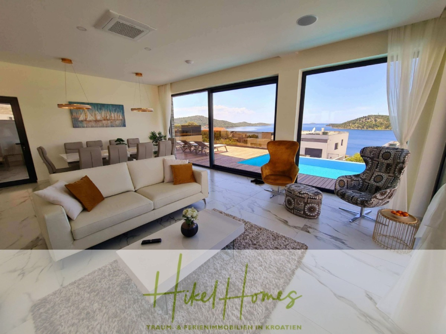 Seeview 2 von 3 | Möbilierte Luxus-Meerblick Villa mit 4 SZ, Pool, Sauna, Fitness, Jacuzzi, - Modernes Wohnzimmer mit weißem Sofa, Stühlen und Essbereich mit großen Fenstern und Blick auf einen Pool, eine Terrasse und eine malerische Landschaft am Wasser in Kroatien. Der Text "Hikel Homes" ist am unteren Rand eingeblendet. (45611)
