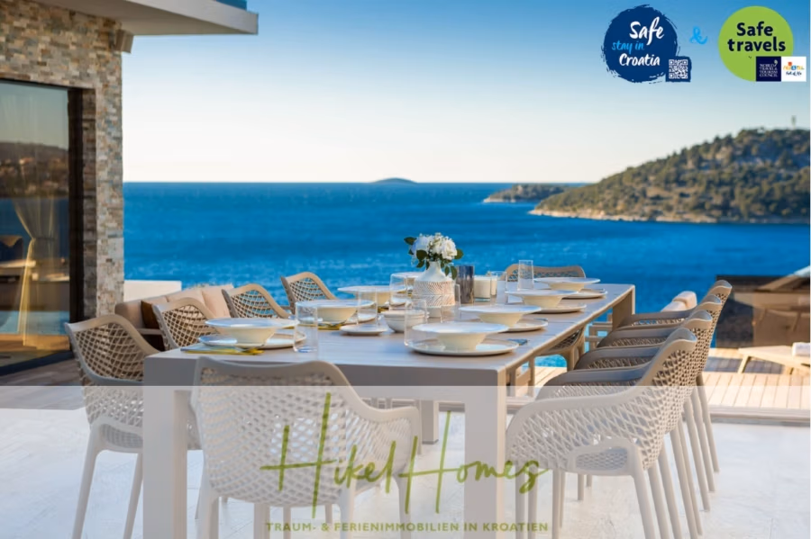 Seeview 2 von 3 | Möbilierte Luxus-Meerblick Villa mit 4 SZ, Pool, Sauna, Fitness, Jacuzzi, - Essbereich im Freien mit Blick auf eine malerische Küste mit klarem, blauem Meer und fernen Inseln. Ein moderner weißer Tisch mit zehn Stühlen ist mit einem Blumenschmuck geschmückt. In der Ecke befinden sich Aufkleber mit der Aufschrift „Sicherer Aufenthalt in Kroatien“ und „Gute Reise“. (41744)