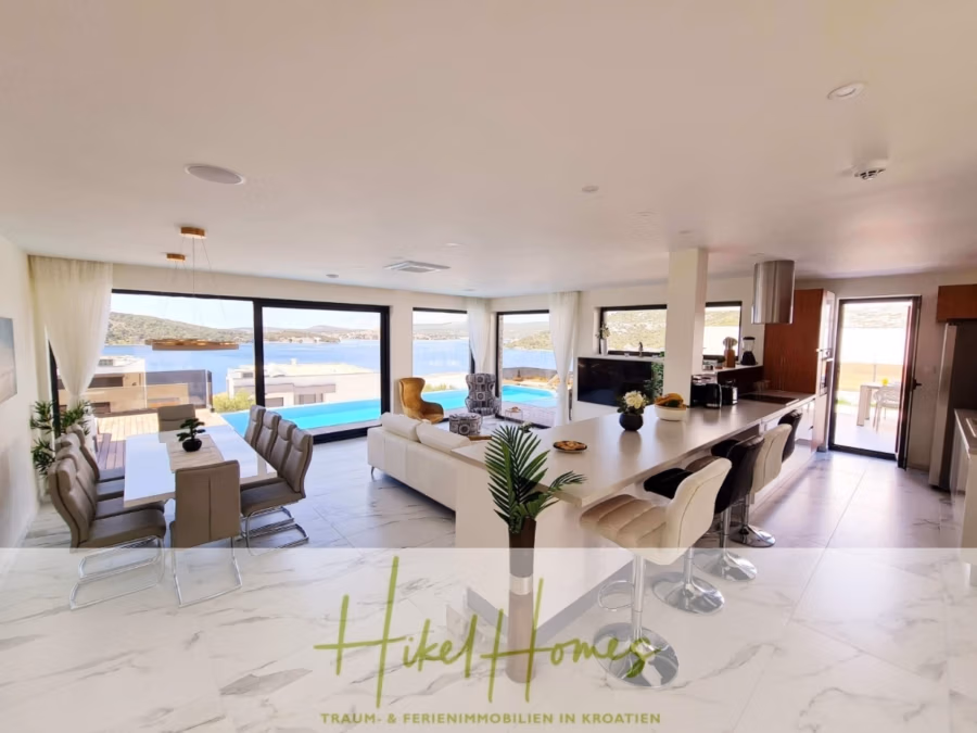 Seeview 2 von 3 | Möbilierte Luxus-Meerblick Villa mit 4 SZ, Pool, Sauna, Fitness, Jacuzzi, - Großzügiger, moderner, offener Wohnbereich mit Esstisch, Sofas und einer Kücheninsel mit Hockern. Große Fenster bieten Blick auf Pool und Meer. Helle Einrichtung in neutralen Tönen und Marmorboden. Unten steht „Hikel Homes“. (41755)