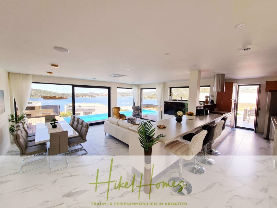 Seeview 2 von 3 | Möbilierte Luxus-Meerblick Villa mit 4 SZ, Pool, Sauna, Fitness, Jacuzzi, - Moderner, offener Wohn- und Essbereich mit großen Fenstern und Blick auf den Pool und die malerische Wasserlandschaft. Zeitgenössisches Mobiliar und neutrale Farben schaffen eine helle, luftige Atmosphäre. Das "Hikel Homes"-Logo ist am Boden angebracht. (45606)