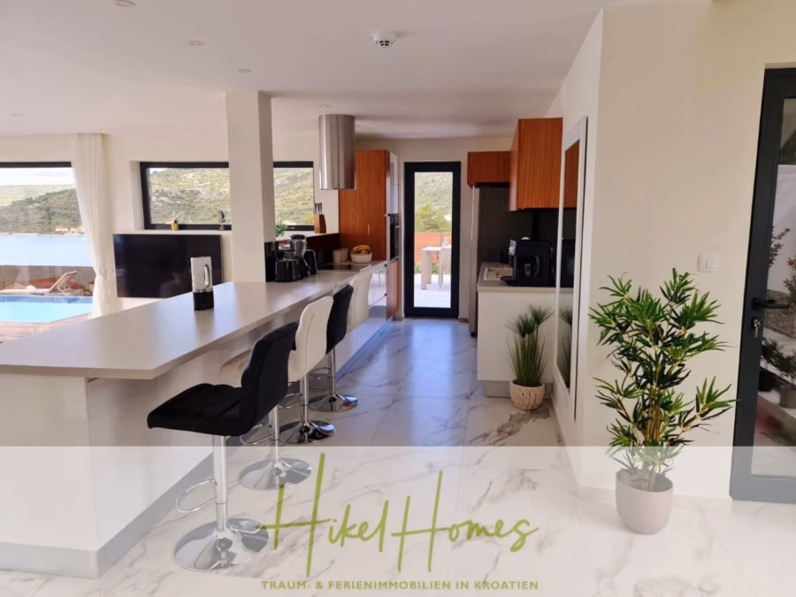 Seeview 2 von 3 | Möbilierte Luxus-Meerblick Villa mit 4 SZ, Pool, Sauna, Fitness, Jacuzzi, - Moderne Küche im eleganten Design mit weißen Arbeitsplatten und dunklen Barhockern. Holzschränke säumen die Rückwand, und ein raumhohes Fenster bietet einen malerischen Ausblick. Eine Topfpflanze verleiht dem Raum einen Hauch von Grün. Logotext unten. (41758)