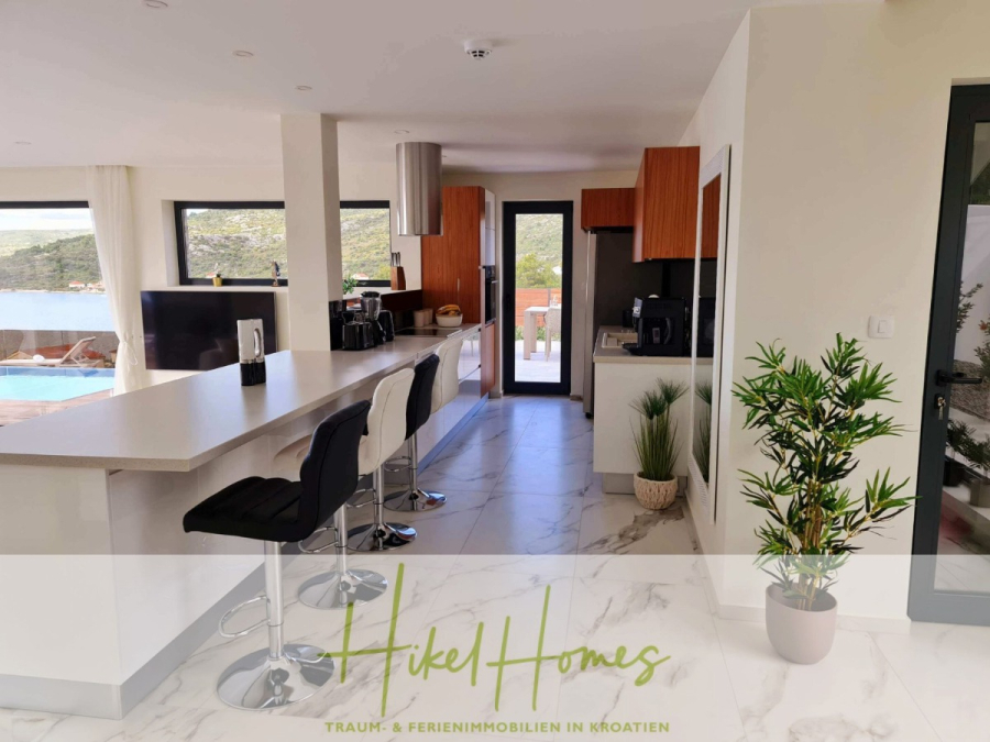 Seeview 2 von 3 | Möbilierte Luxus-Meerblick Villa mit 4 SZ, Pool, Sauna, Fitness, Jacuzzi, - Moderne Küche mit weißen Arbeitsplatten, schwarzen und weißen Barhockern, Holzschränken, Einbaugeräten, großen Fenstern und einer Topfpflanze. Der Pool und die malerische Aussicht nach draußen sind durch die Fenster sichtbar. Der Text lautet: "Hikel Homes. (45610)
