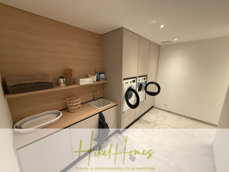 Erstbezug – ab sofort und Herbst genießen! Schlüsselfertige Meerblick Villa, 3SZ, Pool, … - Moderne Waschküche mit Einbauschränken, zwei Frontlader-Waschmaschinen, einem Waschbecken, einer Arbeitsplatte mit Körben, Handtüchern und Reinigungsmitteln, unter warmer Deckenbeleuchtung. (44493)
