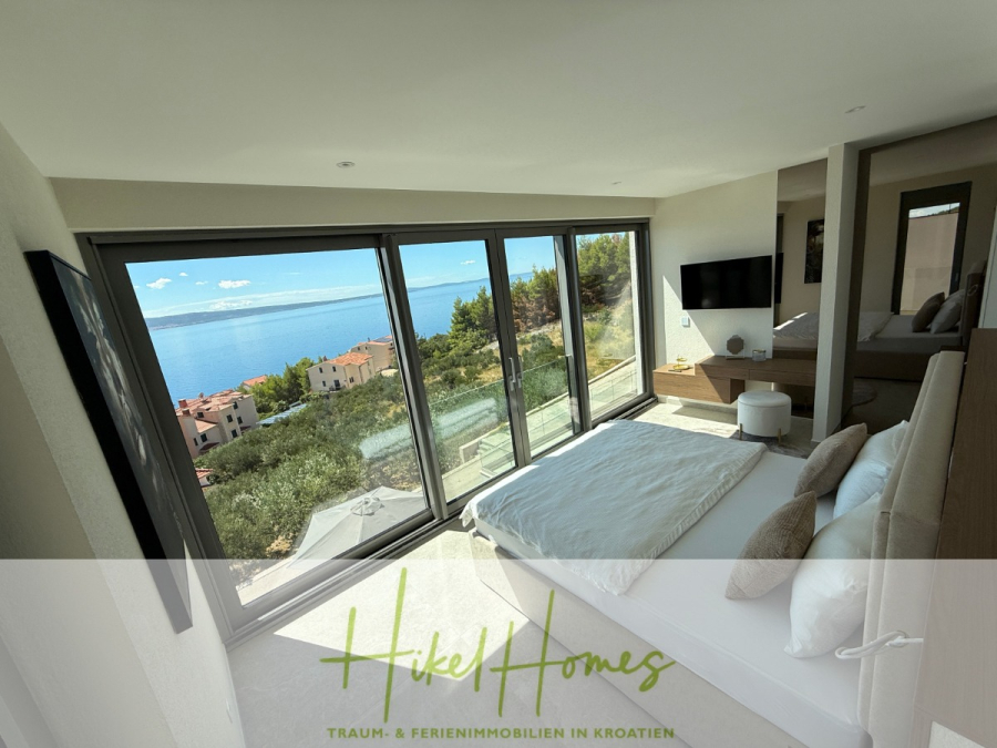 Erstbezug – ab sofort und Herbst genießen! Schlüsselfertige Meerblick Villa, 3SZ, Pool, … - Ein modernes Schlafzimmer mit großen, raumhohen Fenstern bietet einen atemberaubenden Blick auf das Meer, die Hügel und die Küstengebäude. Das Zimmer verfügt über ein ordentlich gemachtes Bett, einen Fernseher und eine minimalistische Einrichtung. Der Text "Hikel Homes" überlagert den Boden. (44480)