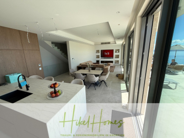 Moderner, offener Wohnbereich mit Kücheninsel, Esstisch und Sofa, mit großen Fenstern und Blick auf eine Terrasse und das Meer. Der Raum ist hell und minimalistisch mit neutralen Tönen. - Erstbezug – ab sofort und Herbst genießen! Schlüsselfertige Meerblick Villa, 3SZ, Pool, …