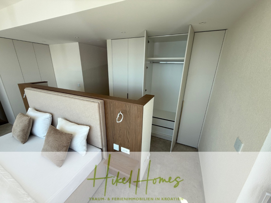 Erstbezug – ab sofort und Herbst genießen! Schlüsselfertige Meerblick Villa, 3SZ, Pool, … - Ein modernes Schlafzimmer mit einem ordentlich gemachten Bett, beigen und braunen Kissen, einem Einbauschrank mit offenen Türen und einem minimalistischen Dekor in Weiß und Holz. Das Logo "Hikel Homes" ist am unteren Rand eingeblendet. (44483)