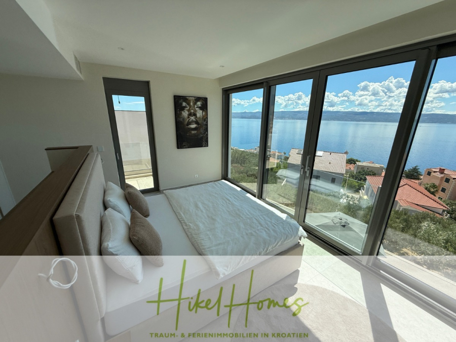 Erstbezug – ab sofort und Herbst genießen! Schlüsselfertige Meerblick Villa, 3SZ, Pool, … - Modernes Schlafzimmer mit großen, bodentiefen Fenstern, die einen malerischen Blick auf das Meer, die Dächer und die fernen Berge bieten. Das Bett steht in der Nähe der Fenster, und ein Porträt hängt an einer weißen Wand. (44484)